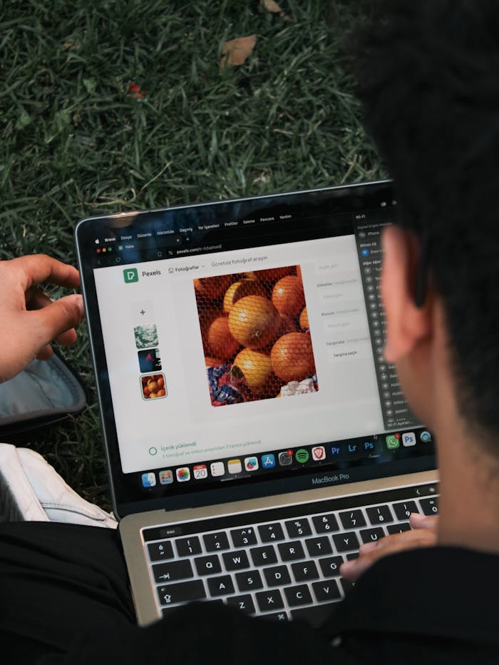 A man edits a fruit image on a MacBook Pro while sitting outdoors on grass.